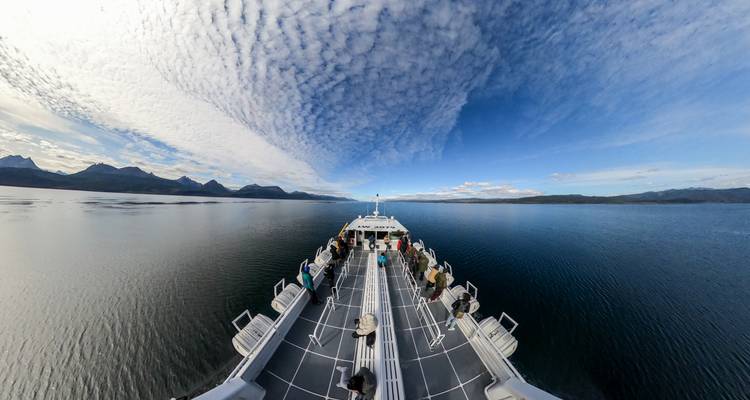Vue en fisheye depuis la proue d'un grand catamaran naviguant sur des eaux bleues calmes sous des nuages aux motifs variés.
