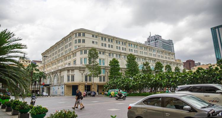 Historisches französisches Kolonialstilgebäude grenzt an einen Platz unter bewölktem Himmel mit vorbeifahrenden Autos und Fußgängern.