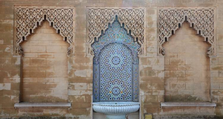 Intricately tiled Moroccan fountain set within an ornate sandstone wall framed by arched niches.