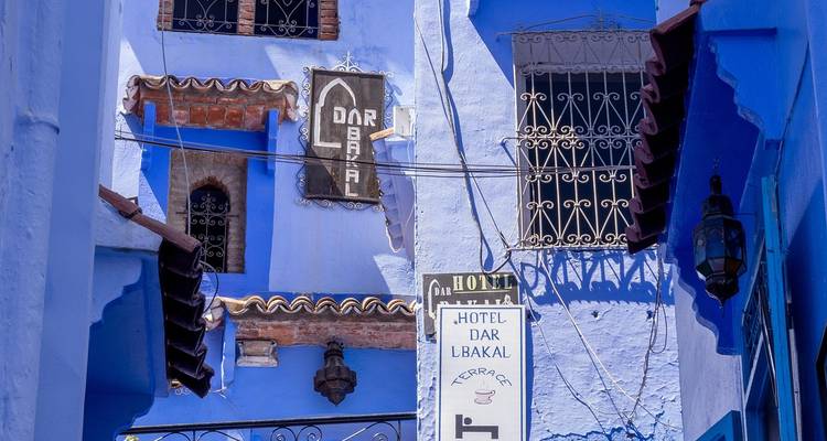 Vibrant blue-painted alley in Chefchaouen with decorative signs, wrought-iron windows and hanging cables.