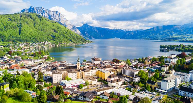 Luchtfoto van een Oostenrijkse stad aan een meer, omringd door steile groene bergen en een breed alpenmeer onder verspreide wolken.