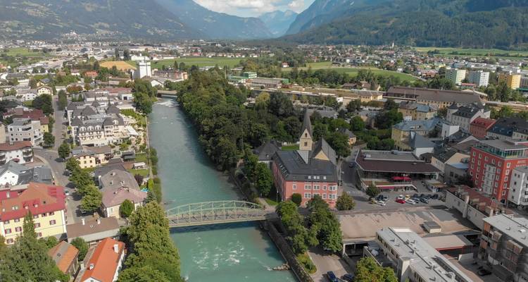 Dronebeeld van een turquoise rivier die door een Tiroolse stad stroomt, omringd door alpentoppen en groene velden.