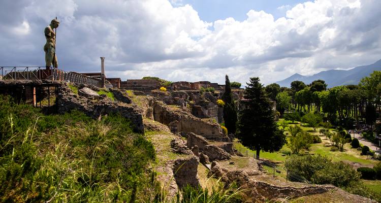 Terrazas de piedra en ruinas y una gran estatua de bronce que domina el sitio arqueológico con montañas al fondo.