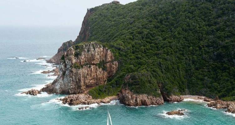 Un voilier solitaire navigue dans des eaux turquoise entre des falaises maritimes spectaculaires couvertes de végétation.