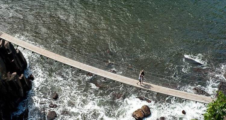 Passerelle suspendue traversant des eaux de marée écumantes avec un marcheur solitaire capturé d'en haut.