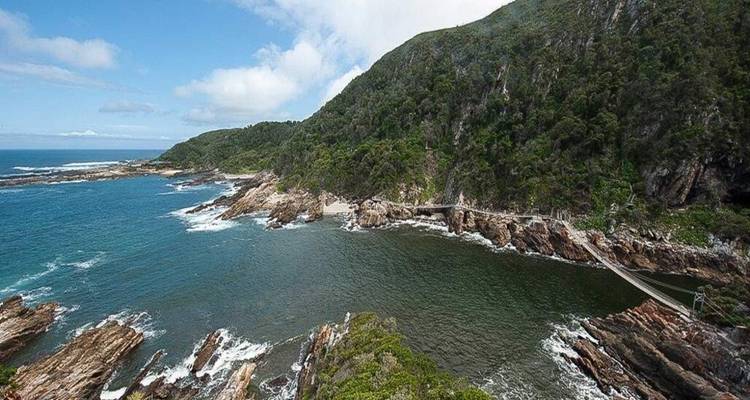 Vue large d'une gorge boisée où un pont suspendu enjambe une crique tranquille qui rejoint la mer.