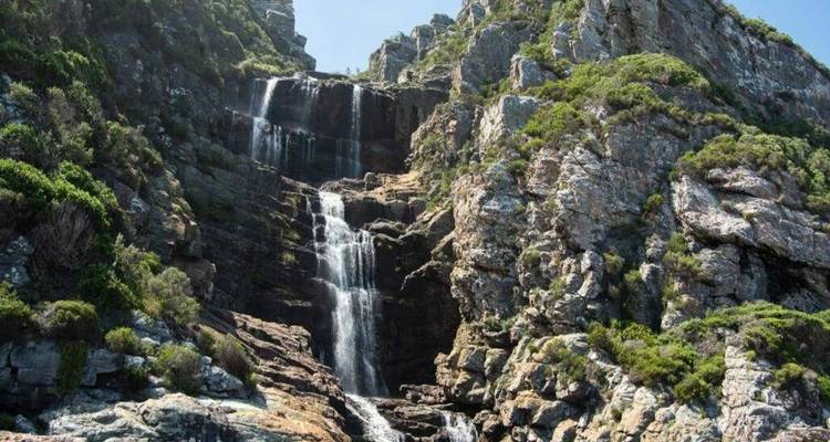 Cascade à plusieurs niveaux dévalant des formations rocheuses escarpées entourées d'arbustes vert vif.