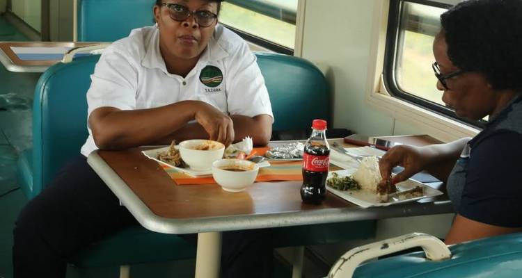 Two women sit in a train dining car eating local food with a soda bottle on the table.