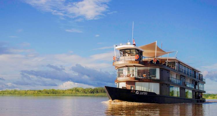 Un bateau de croisière fluviale moderne à plusieurs ponts glisse le long d'une large rivière de jungle sous un ciel bleu.