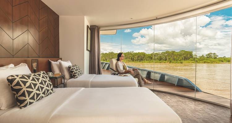 Cabine spacieuse à lits jumeaux avec baie vitrée du sol au plafond donnant sur une rivière de forêt tropicale où une femme se détend près de la fenêtre.