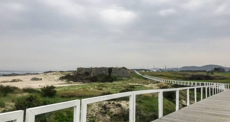 Lange houten promenade loopt langs duinen en een oud stenen fort onder grijze luchten.