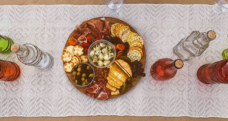 Bovenaanzicht van een gourmet charcuterieplank met vleeswaren, kazen, olijven en crackers geflankeerd door gekleurde wijnflessen.
