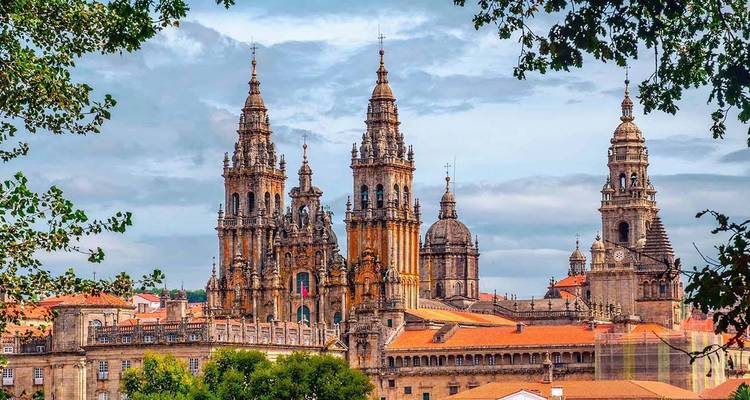 Iconische kathedraal van Santiago de Compostela met twee torens omlijst door groene takken tegen een gedeeltelijk bewolkte hemel.