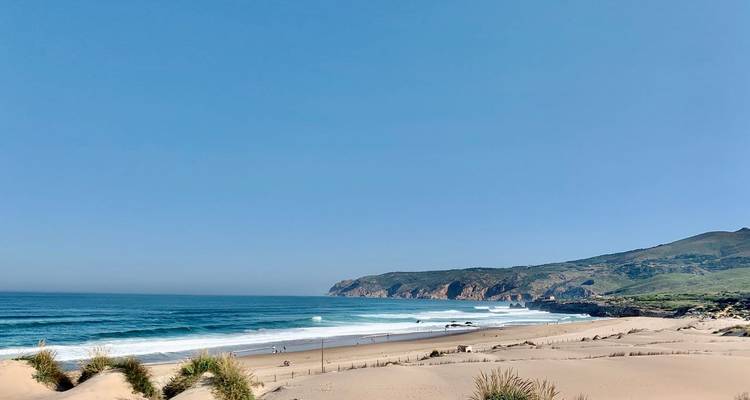 Breiter Sandstrand mit rollender Brandung unter einem klaren blauen Himmel.