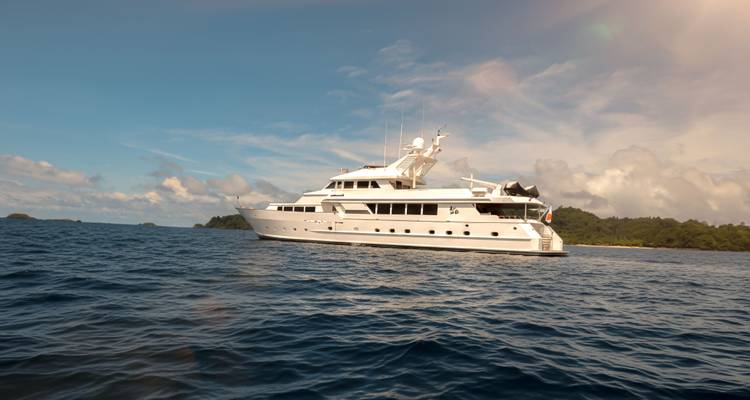 Grand yacht de luxe blanc amarré en pleine mer avec des îles verdoyantes luxuriantes en arrière-plan sous un ciel bleu.