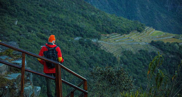 Un randonneur en veste rouge et bonnet orange observe des ruines en terrasses dans une vallée andine luxuriante.