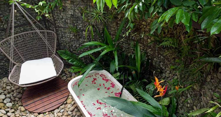 Vrijstaand buitenbad bestrooid met bloemblaadjes naast hangende stoel in bladrijke hoek.