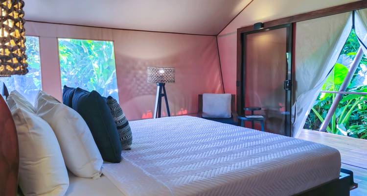 Lichte glamping slaapkamer met kingsize bed, gekussens met patronen en uitzicht op privé terras en groen.