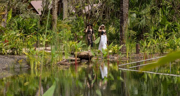 Twee reizigers in resortkleding staan naast een reflecterende junglevijver te midden van dichte begroeiing.