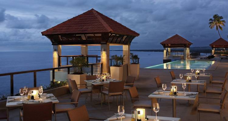 Elegant open-air restaurant terrace beside an infinity pool overlooking the sea at dusk, tables set with wine glasses and candles, dramatic cloudy sky beyond.