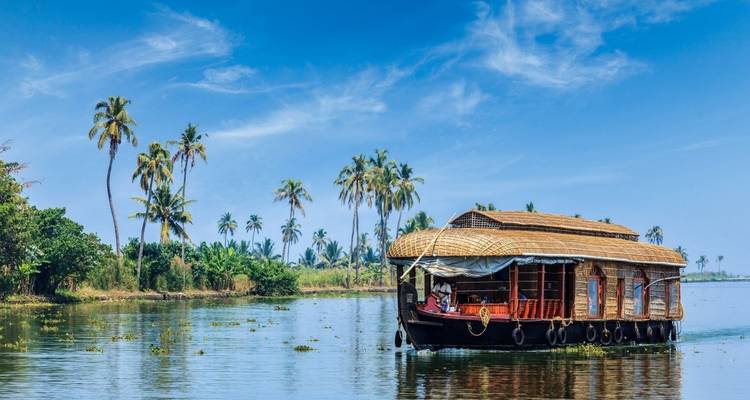 Traditional thatched-roof Kerala houseboat cruising the lush green backwaters bordered by palm trees under a clear blue sky.
