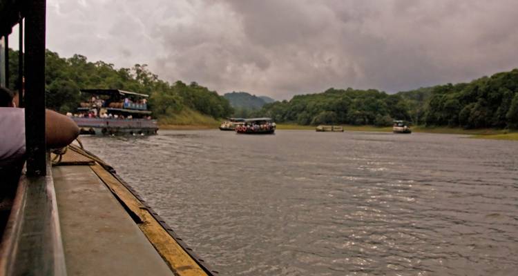 View from inside a tour boat on a wide lake with several other passenger boats ahead under a cloudy sky and surrounding green hills.