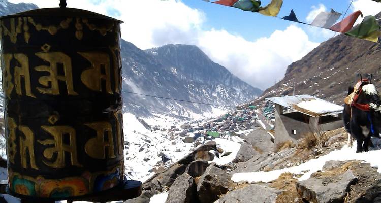 Großes Gebetsrad im Vordergrund mit goldener Schrift, schneebedecktes Tal und entferntes Bergdorf dahinter, Yak an der Seite.