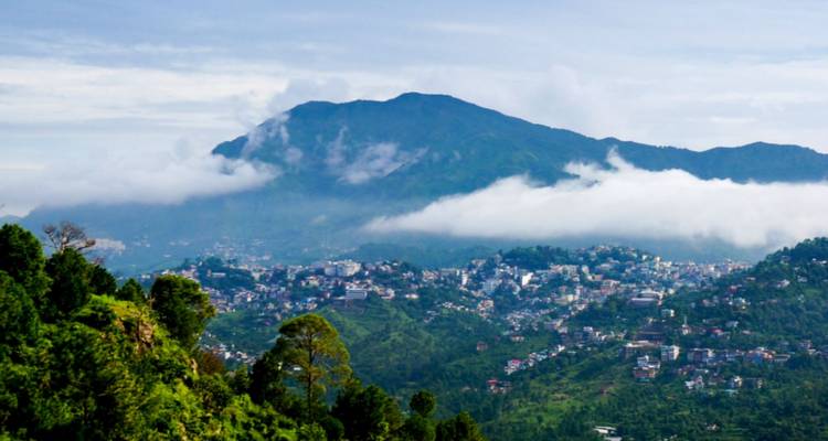 Ausgedehnte Bergkulisse über einer weitläufigen Hügelstadt, die teilweise von tiefen Wolken verschleiert und von Wald umgeben ist.