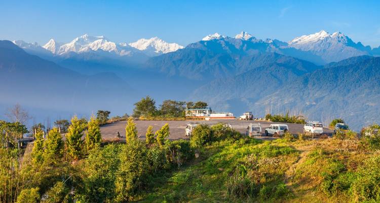 Hohe Aussicht auf die schneebedeckte Kanchenjunga-Kette über einer Bergstraße mit geparkten Touristenbussen und üppigem Grün.