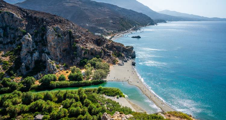 Luftaufnahme des Preveli-Strandes, wo ein grüner Fluss inmitten von Palmenwäldern und schroffen Bergen auf das Meer trifft.