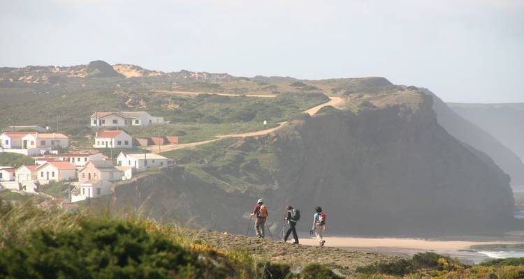 Drie wandelaars met rugzakken lopen langs een kustpad boven een zandige baai en witte huizen met dramatische kliffen erachter