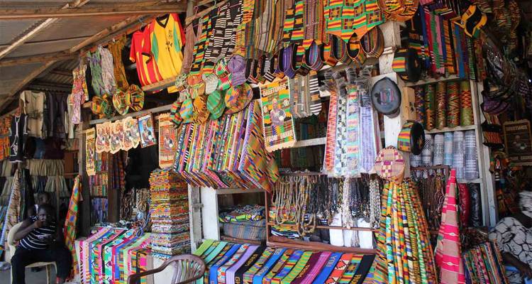 Bustling craft market stall displaying brightly coloured kente fabrics and beaded jewellery.