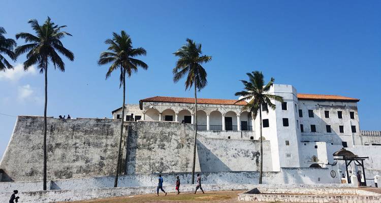 Historic white-washed Elmina Castle with tall palms and blue sky towering above coastal ramparts.