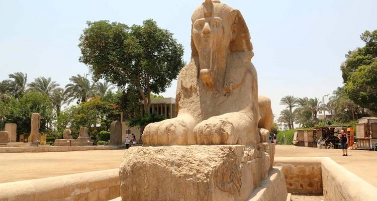 Ancient alabaster sphinx statue in open archaeological garden with palms behind.