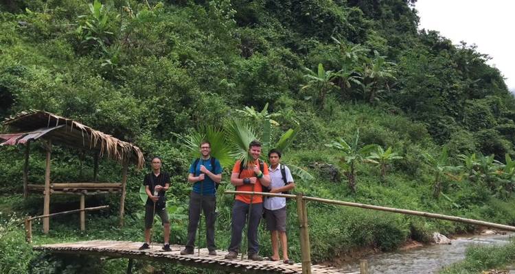 Kleine Gruppe steht auf einer rustikalen Bambusbrücke über einem klaren Bach mit Dschungel-Kulisse.