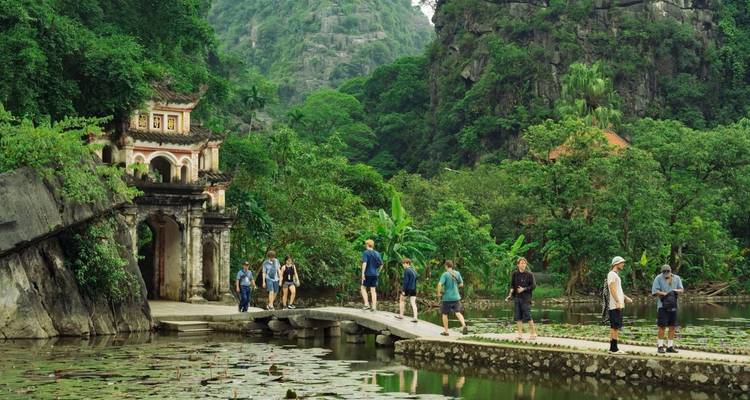 Los visitantes caminan por una calzada de piedra junto a un estanque de loto hacia un templo ornamentado situado entre acantilados de piedra caliza exuberante.