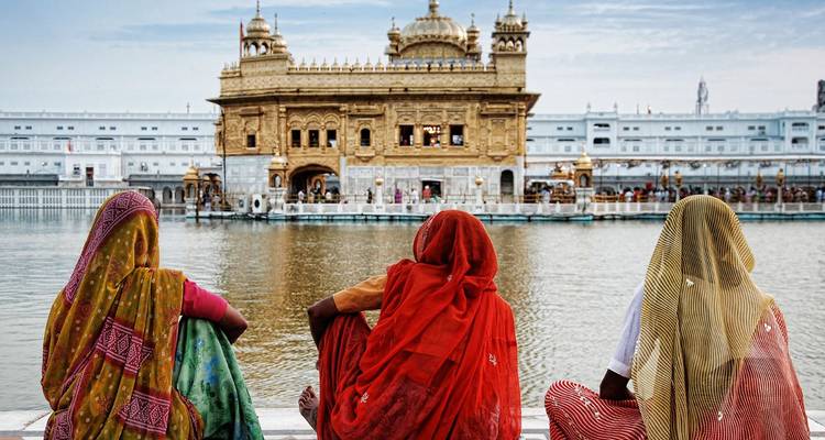 Tres mujeres con saris coloridos se sientan junto a una piscina reflectante mirando hacia el Templo Dorado al otro lado del agua.