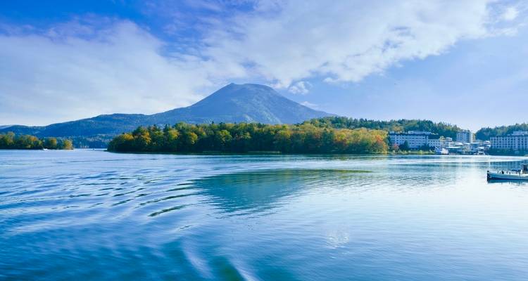 Un lac bleu calme reflète des collines boisées avec un sommet lointain sous un ciel matinal lumineux