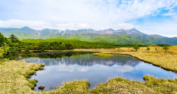 L'étang immobile comme un miroir reflète la forêt verdoyante et les pics neigeux lointains sous un ciel bleu