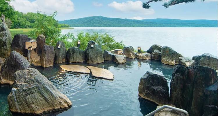 Piscine naturelle de source chaude en plein air bordée de roches avec vue sur un lac serein et des collines boisées.