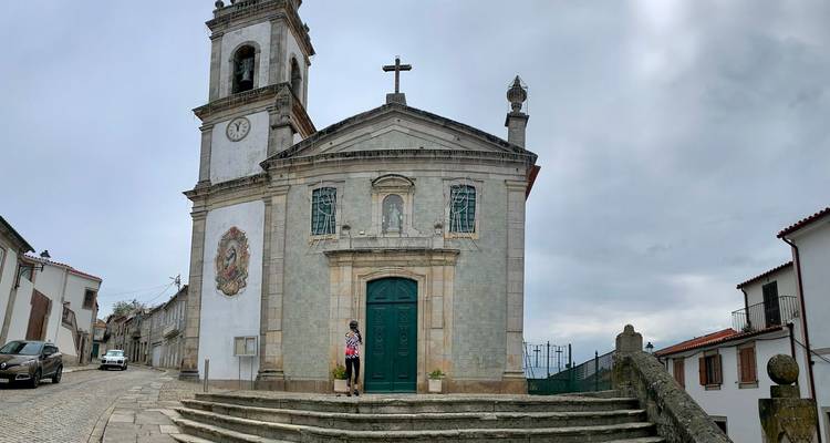 Historische Steinkirche mit Glockenturm und Kreuz steht auf einem Kopfsteinpflasterplatz unter bewölktem Himmel, einzelner Besucher an der Tür