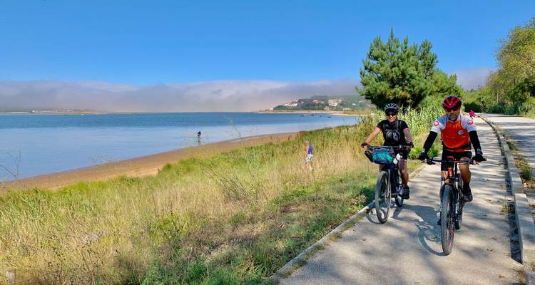 Zwei Radfahrer, die auf einem asphaltierten Weg am Seeufer fahren, mit nebligen Hügeln im Hintergrund