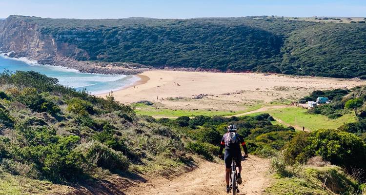 Radfahrer fährt einen Feldweg hinunter zu einer abgelegenen Sandbucht, umgeben von grünen Klippen