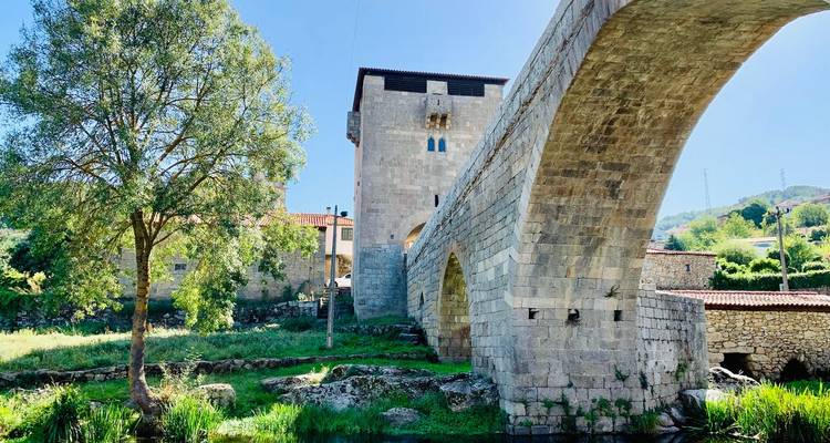 Steinerne mittelalterliche Brücke mit Turm überspannt einen bewachsenen Bach, gerahmt von blauem Himmel