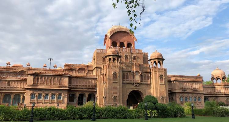 De sierlijke roze zandstenen gevel van Laxmi Niwas Palace tegen keurig onderhouden gazons en een gedeeltelijk bewolkte hemel.