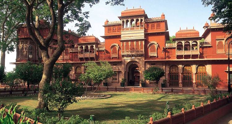 La fachada de arenisca roja y los patios de un palacio rajput están sombreados por grandes árboles y césped bien cuidado.