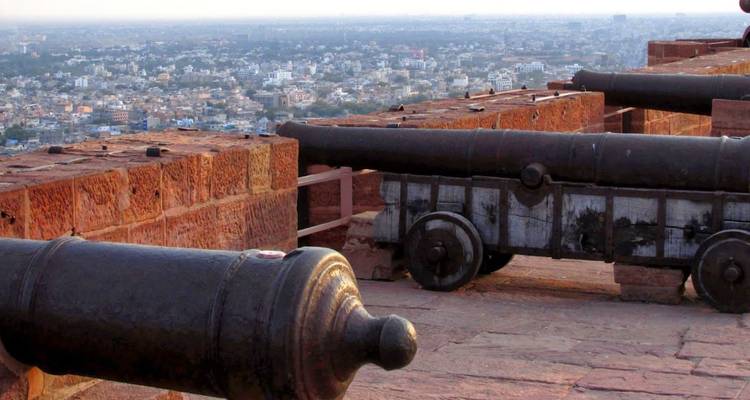 Viejos cañones bordean las murallas de un fuerte en la cima de una colina con vista a una extensa ciudad india.