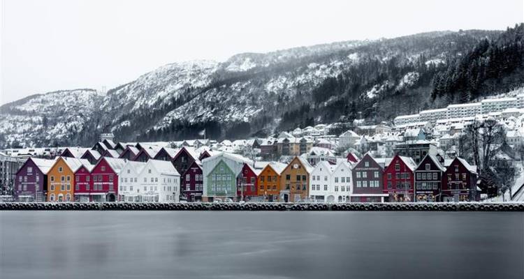 Reihe von bunt bemalten hölzernen Bryggen-Lagerhäusern, die sich in einem ruhigen, schneereichen Fjord mit kiefernbewaldeten Bergen im Hintergrund spiegeln