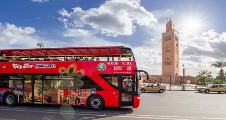 Un autobús turístico urbano rojo descapotable pasa frente a la Mezquita Kutubía en una soleada calle de Marrakech.