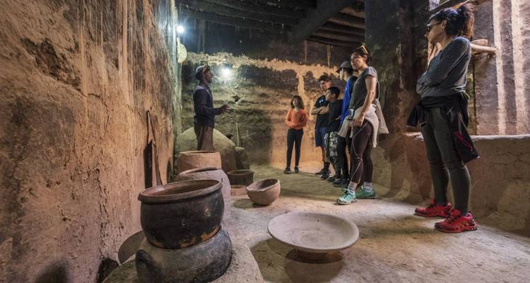 Un pequeño grupo escucha a un guía dentro de una casa histórica de paredes de tierra llena de cerámica antigua.